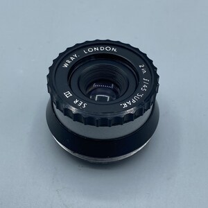 Pu&ograve; includere: Un obiettivo nero Wray London Super 2in f/4.5 con un anello argentato e il testo "Super 2in f/4.5" e "Wray London" sul lato.