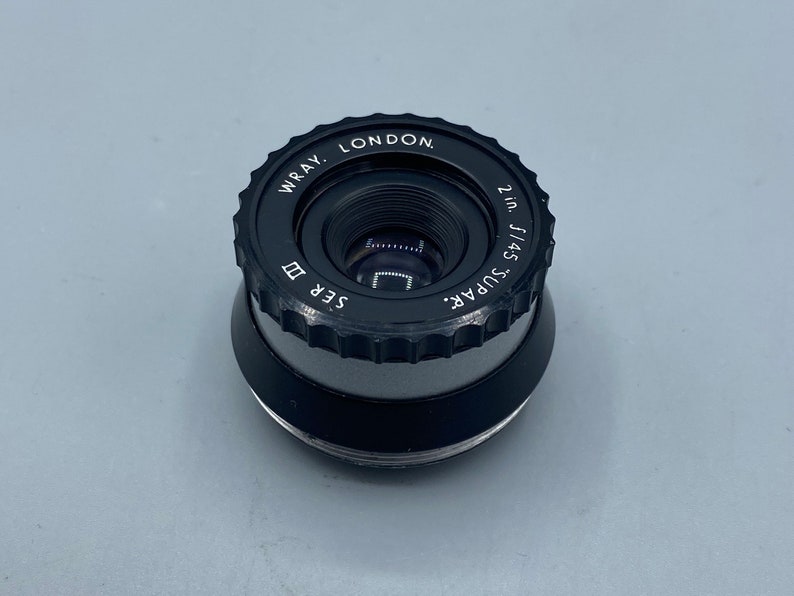 Pu&ograve; includere: Un obiettivo nero Wray London Super 2in f/4.5 con un anello argentato e il testo "Super 2in f/4.5" e "Wray London" sul lato.