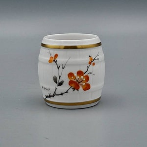 Puede incluir: Un pequeño recipiente de cerámica blanca en forma de barril con un borde dorado y un diseño floral con flores naranjas y ramas negras.