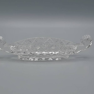 May include: A clear glass serving dish with a decorative cut-glass pattern. The dish has two handles on either side and a rounded base.