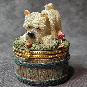 May include: A decorative trinket box featuring a cream-colored dog figurine with a playful pose. The box is barrel-shaped, with a wooden texture and blue accents. A ladybug and yarn balls add detail to the scene.