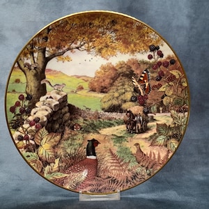 May include: Decorative plate featuring a pastoral scene with a pheasant, a butterfly, and a horse-drawn cart. The plate has a gold rim and depicts a stone wall, trees, and a field. The colors are warm, with browns, greens, and touches of red.