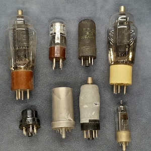Może przedstawiać: Kolekcja zabytkowych lamp próżniowych, z szklanymi bańkami o różnych kształtach i podstawach. Niektóre lampy mają brązowe, beżowe lub czarne podstawy. Lampy są ułożone na szarej powierzchni, prezentując swoje wewnętrzne elementy.