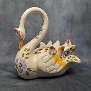 May include: A ceramic swan ornament with a curved neck and open back. The swan is white with yellow, blue, and green floral accents. The beak is yellow and the head has brown accents. The swan is a decorative item.