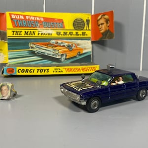 Puede incluir: Un coche de juguete de metal azul con un techo blanco y una luz plateada en la parte superior. El coche tiene una parrilla negra y ruedas negras. El coche está etiquetado como "THRUSH-BUSTER" y "GUN FIRING". El coche es de la serie "El hombre de U.N.C.L.E.". El coche está sentado en una mesa con una caja detrás. La caja es amarilla y tiene una imagen del coche en ella. La caja también tiene las palabras "GUN FIRING THRUSH-BUSTER" y "THE MAN FROM U.N.C.L.E." en ella.