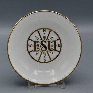 May include: A white ceramic dish with a gold rim and a compass design in the center. The compass features the letters "ESU" in a stylized font. The dish is displayed on a clear plastic stand.