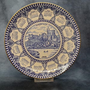 May include: A decorative plate featuring a blue and white design with a gold rim. The plate has a calendar around the edge and a central image of Windsor Castle with the year 1984.