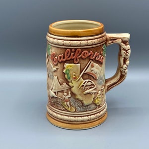 Può includere: Un boccale di birra in ceramica con manico, con un design a rilievo dei monumenti della California. Il boccale è marrone e marrone con la scritta "California" in rosso. Il manico ha un design a vite.