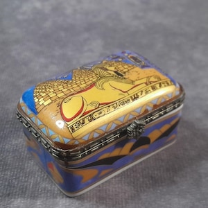 May include: A rectangular trinket box with a hinged lid, featuring an Egyptian theme. The box is primarily blue, gold, and white, with an illustration of a sphinx and pyramids. The box has a metal frame and clasp.
