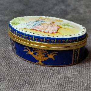 Può includere: Scatola portagioie ovale con base blu e bordo dorato. Il coperchio presenta una scena dipinta con figure in un giardino. La scatola ha un design decorativo dorato sul davanti.