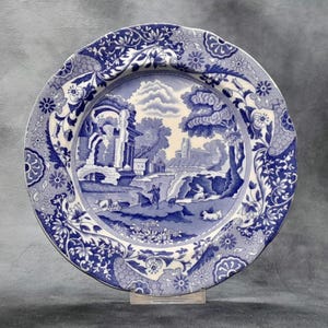 Puede incluir: Un plato decorativo azul y blanco con una escena de paisaje. El plato presenta un diseño detallado de una escena pastoral con edificios, árboles y figuras. El borde tiene un patrón floral y de desplazamiento. El plato se exhibe sobre un soporte transparente.