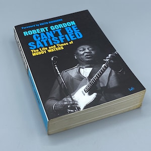 May include: A black and blue book titled "Can't Be Satisfied: The Life and Times of Muddy Waters" by Robert Gordon with a foreword by Keith Richards. The cover features a black and white photo of Muddy Waters playing a guitar.