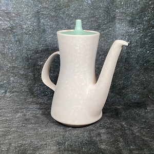 Puede incluir: Una cafetera de cerámica blanca con una tapa azul claro. La cafetera tiene una superficie texturizada y un pico curvo.