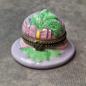 May include: A small, decorative hat-shaped trinket box. The box is primarily lavender with pink and green accents. The lid features a green leaf design and a brass-colored metal trim. The base is round and the box is likely ceramic.