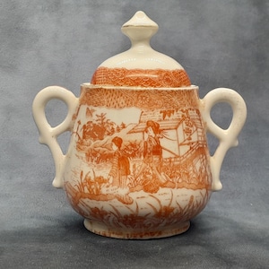 Puede incluir: Azucarero de cerámica color crema con tapa, con un diseño naranja de transferencia de un paisaje con figuras. El azucarero tiene dos asas y un adorno decorativo en la tapa. Una pieza vintage.
