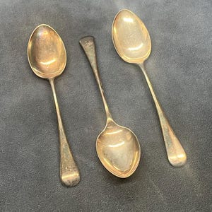Op de afbeelding: Drie antieke zilveren lepels met ovale bakken en lange, slanke stelen. Eén lepel heeft een initiaal op de steel. De lepels zijn gerangschikt op een donkergrijze ondergrond, wat hun verouderde, aangetaste uiterlijk laat zien.