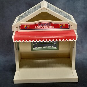 May include: A vintage toy shop with a red awning and the words "GAMES SOUVENIRS" on a sign. The shop has a cream-colored exterior, a patterned roof, and a small window. The toy shop is a miniature replica.