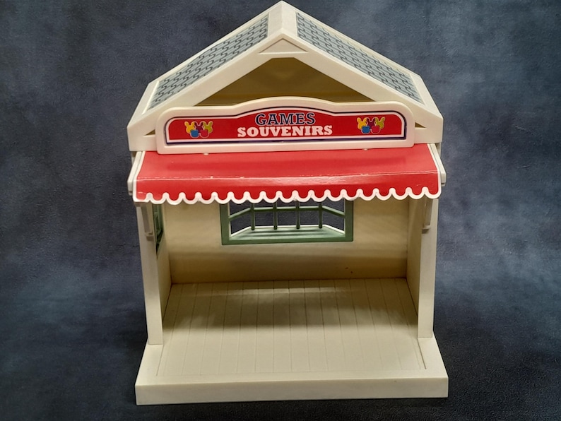 May include: A vintage toy shop with a red awning and the words "GAMES SOUVENIRS" on a sign. The shop has a cream-colored exterior, a patterned roof, and a small window. The toy shop is a miniature replica.