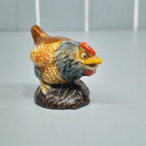 Puede incluir: Una figurita de cerámica de un pollo marrón y azul con una cresta roja y un pico amarillo. El pollo está de pie sobre una base marrón.