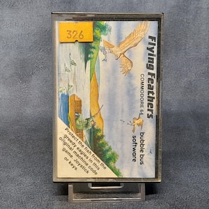 Puede incluir: Un cartucho de juego Commodore 64 vintage para "Flying Feathers". La etiqueta presenta una ilustración de águilas, un pescador y el texto "Protect the fish from the greedy eagles". Una pegatina amarilla muestra el número "326".