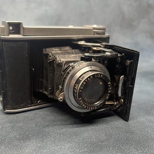 Puede incluir: Una cámara Voigtlander vintage en blanco y negro con una lente abierta. La lente está marcada "Voigtar 1:3.5 F=7.5 cm". La cámara está sobre una superficie gris.