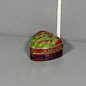 Puede incluir: Una pequeña caja de joyas triangular con una base roja y un borde azul y dorado. La tapa está decorada con un diseño floral verde y dorado.