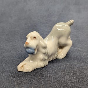 Puede incluir: Una pequeña figura de cerámica blanca brillante de un perro en una posición juguetona y agachada. El perro tiene orejas largas y texturizadas y sostiene una pelota redonda, lisa y de color azul claro en la boca. Acabado esmaltado.