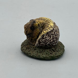 May include: A small, curled-up hedgehog figurine with brown and gold fur, sitting on a green base.