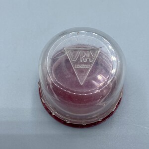 Pu&ograve; includere: Un contenitore di plastica trasparente con una base rossa e un logo triangolare argentato che dice "VRAY LONDON".