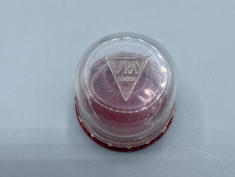 Pu&ograve; includere: Un contenitore di plastica trasparente con una base rossa e un logo triangolare argentato che dice "VRAY LONDON".