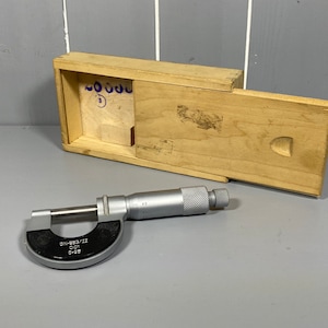 Könnte beinhalten: Ein silberner Mikrometer mit einem schwarzen Zifferblatt und einer Holzkiste. Das Zifferblatt zeigt "DN-683/II 0.01 0-25".