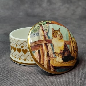 Puede incluir: Pequeña caja de baratijas de cerámica blanca abierta con tapa decorativa. La tapa presenta una ilustración detallada de un gato sentado en una silla. El exterior de la caja está adornado con corazones, estrellas y diamantes dorados.