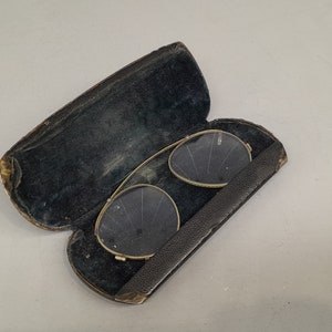 May include: A pair of gold-rimmed glasses with round lenses in a black leather case. The case is open and the glasses are visible inside.