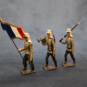 May include: Three miniature toy soldiers in tan uniforms, white hats, and black boots. One carries a French flag, another a bugle, and the third a rifle. The figures stand on rectangular bases against a gray background.