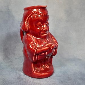 May include: A red ceramic jug shaped like a person wearing a robe. The jug has a handle and a spout.