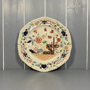 Puede incluir: Un plato de cerámica blanco con un patrón floral decorativo en tonos de azul, rojo, naranja y verde. El plato presenta un diseño central de una valla estilizada con un motivo floral.