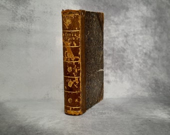 Op de afbeelding: Een antiek boek met een versleten, bruine leren kaft en gouden accenten. De rug heeft reliëftekst en decoratieve elementen. De pagina's zijn verouderd en de randen vertonen slijtage, wat wijst op de leeftijd en het gebruik ervan.
