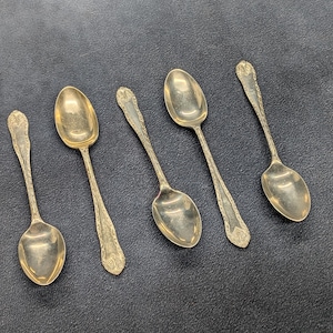 May include: A set of five silver-toned metal spoons arranged in a row. Each spoon features an elongated oval bowl and a decorative handle with intricate embossed detailing at the top and base, showcasing a tarnished metallic finish throughout.