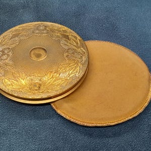 May include: A vintage gold-toned compact with an ornate floral design on the lid. The compact is open, revealing a round, tan powder puff. The compact is on a blue surface.