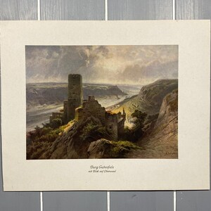 Op de afbeelding: Een vintage print van een schilderij dat een kasteelruïne op een heuveltop toont met uitzicht op een rivier. Het kasteel is van steen en heeft een hoge toren. De rivier is breed en stroomt door een vallei. De lucht is bewolkt en de zon schijnt.