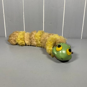 Könnte beinhalten: Ein grüner Plüschfrosch mit gelben Augen und einem gelben und braunen, flauschigen Körper. Der Frosch liegt auf der Seite mit dem Kopf nach rechts gedreht.