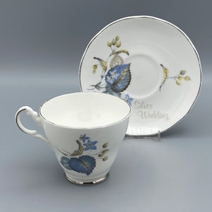 Puede incluir: Una taza de té y un platillo blancos con un diseño floral azul y verde. El platillo tiene las palabras "Silver Wedding" impresas.