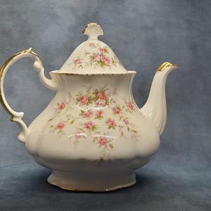 Peut inclure: Théière en porcelaine blanche ornée de motifs floraux roses et verts. La théière présente des détails dorés sur la poignée, le bec et le couvercle. Le couvercle est surmonté d'un bouton décoratif. Un design classique pour servir le thé.