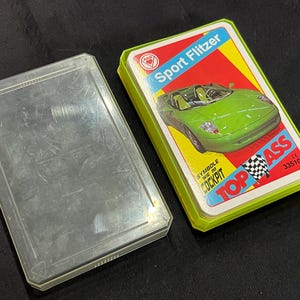 Puede incluir: Una baraja de cartas Sport Flitzer en una caja verde y amarilla. El diseño de la carta presenta un coche deportivo verde con el texto "Sport Flitzer" y "Top Ass". La caja de plástico transparente está abierta.