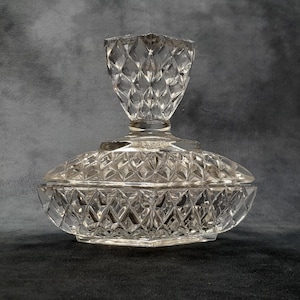 May include: A clear, vintage glass candy dish with a diamond-cut pattern. The dish has a lid with a decorative finial and a rounded base. The glass reflects light, creating a sparkling effect. A classic piece for display or serving.