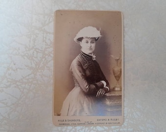 May include: A black and white portrait of a woman wearing a white hat with a feather. She is wearing a dark dress with a lace collar and is standing in front of a vase.
