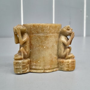 May include: A small, cylindrical stone vase with two carved monkey figures on either side. The vase is a light brown color and has a smooth, polished finish.