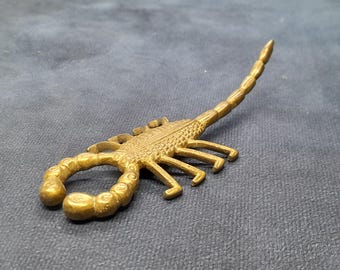 May include: A detailed, golden-colored scorpion figurine. The scorpion has a textured body, curved tail, and multiple legs. The figurine is set against a dark gray background, highlighting its intricate design and metallic finish.