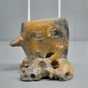 May include: A small, carved stone sculpture of a stylized animal with a yellow and black color scheme. The sculpture has a hole in the top and sits on a base with a rough, textured surface. The sculpture has the Chinese characters "王王" carved into its side.