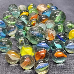 Peut inclure: Un ensemble de billes en verre transparent avec des tourbillons et des motifs colorés. Les billes présentent des nuances de bleu, d'orange, de jaune et de vert. Certaines billes ont un tourbillon blanc laiteux. Les billes varient en taille.